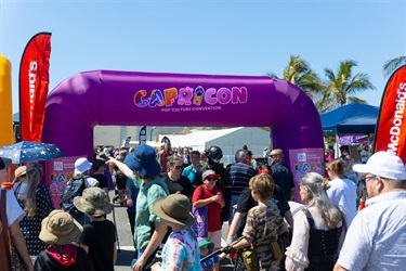 A crowd of people dressed in casual clothes and costumes walk under an inflated purple arch reading 'CapriCon - Pop Culture Convention'