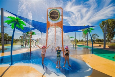 Three children are being splashed by water poured from a large bucket held up by a colourful structure at a water play area.
