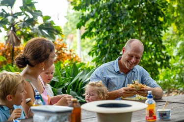 A father smiles as he eats a burger while sitting at a picnic table with his wife and children