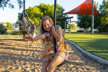 A young girl waves at the camera as she plays on a flying fox and her family watch from the background