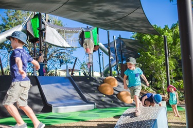 Two children are running across play-equipment ramps, in front of a large playground with slides, climbing walls and rope climbs