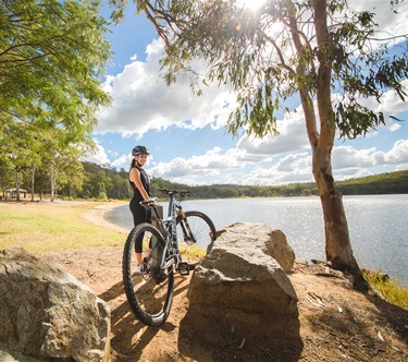 A young woman wearing a helmet stands behind a mountain bike. A lake and green picnic area sits behind her