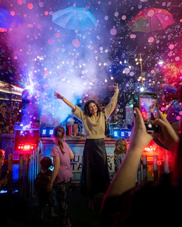 A woman raises her arms in the air in celebration as bubbles blow into the air behind her, lit in red and blue from spotlights. Another person is taking her photo using a mobile phone camera