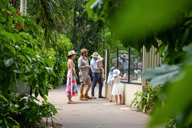A young family looks in on a meerkat enclosure speaking to a zoo-keeper along a tree-lined path
