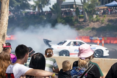 Seated couples, families and children are watching a modified car drifting on a designated arena space