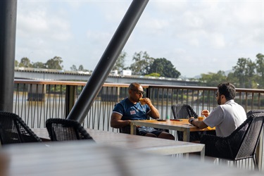 Two men sit on a balcony cafe drinking coffees, overlooking a river. Cars pass over the Fitzroy River Bridge in the background.