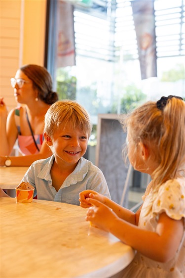 A young boy and girl look at each other smiling, while sitting at a table and eating ice-cream