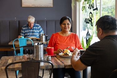 A couple is sitting at a window-side table in a cafe, drinking coffees and eating eggs on toast. She is looking out the window at a tree-lined street