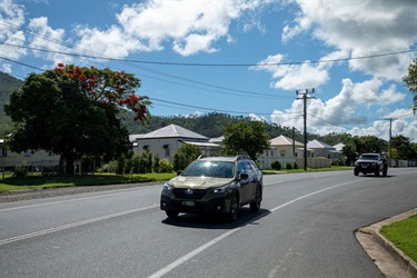 Two cars pass by a row of cottage-style houses with mountains visible in the distance