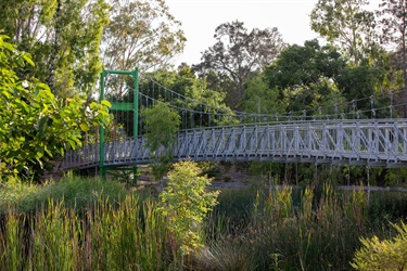 A pedestrian suspension bridge crosses over a lagoon in a well vegetated park