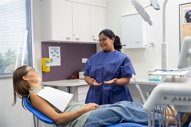 A woman wearing scrubs speaks to a patient reclined on a chair inside a dental clinic
