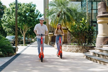 A young man and woman ride down a stone-paved footpath on orange e-scooters, beside a historic building and garden
