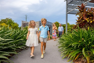 A young boy and girl hold hands as they walk along the footpath leading to a shopping centre, while their parents and sister follow behind
