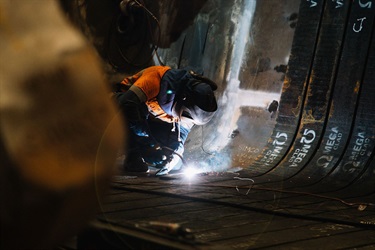 A man wearing protective clothing and a welding mask is squatting on a piece of metal, using a welding torch to join several other strips of metal. Sparks are coming from the end of the welding torch