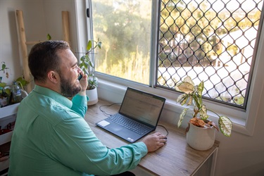 A man sits at a desk and talks on the phone, looking out a window overlooking Rockhampton
