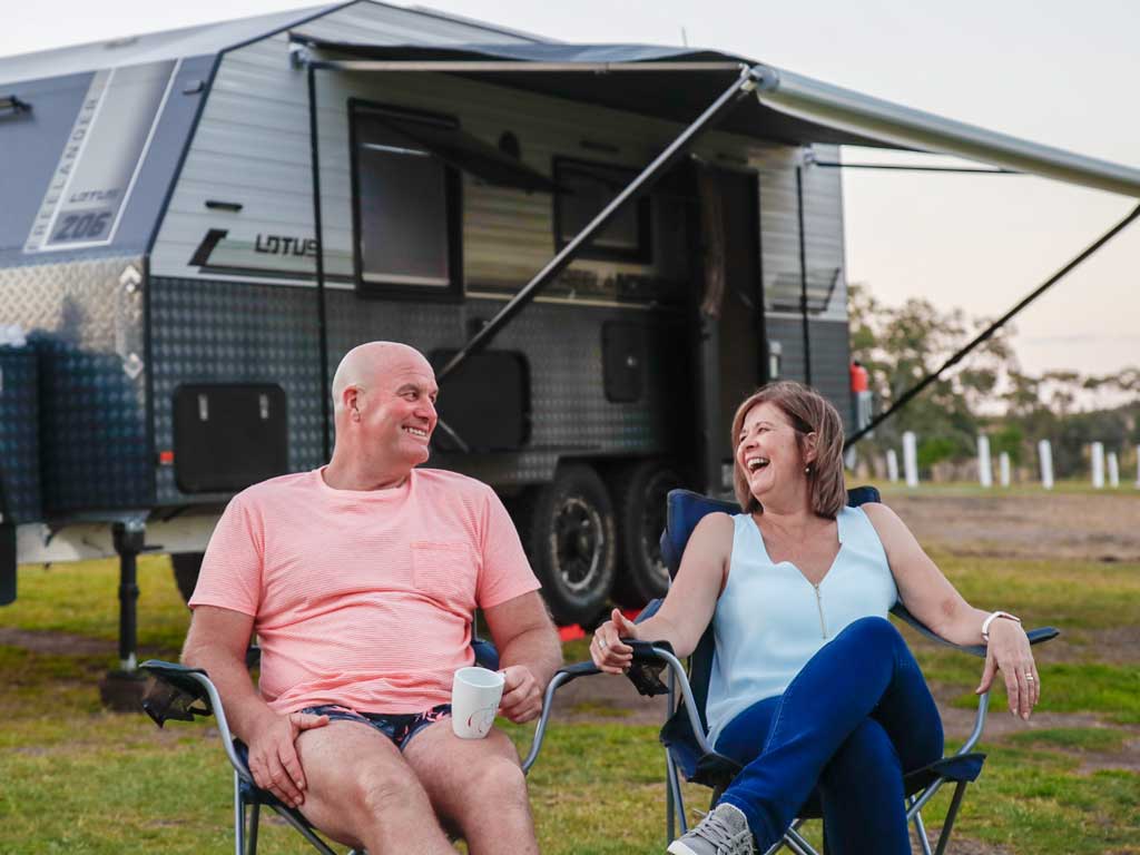 A man and woman sit on camp chairs in front of a parked up caravan