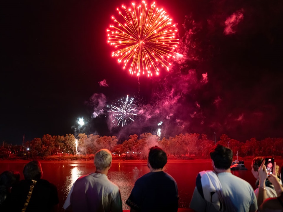 A row of people watch red fireworks light up the sky above the Fitzroy River