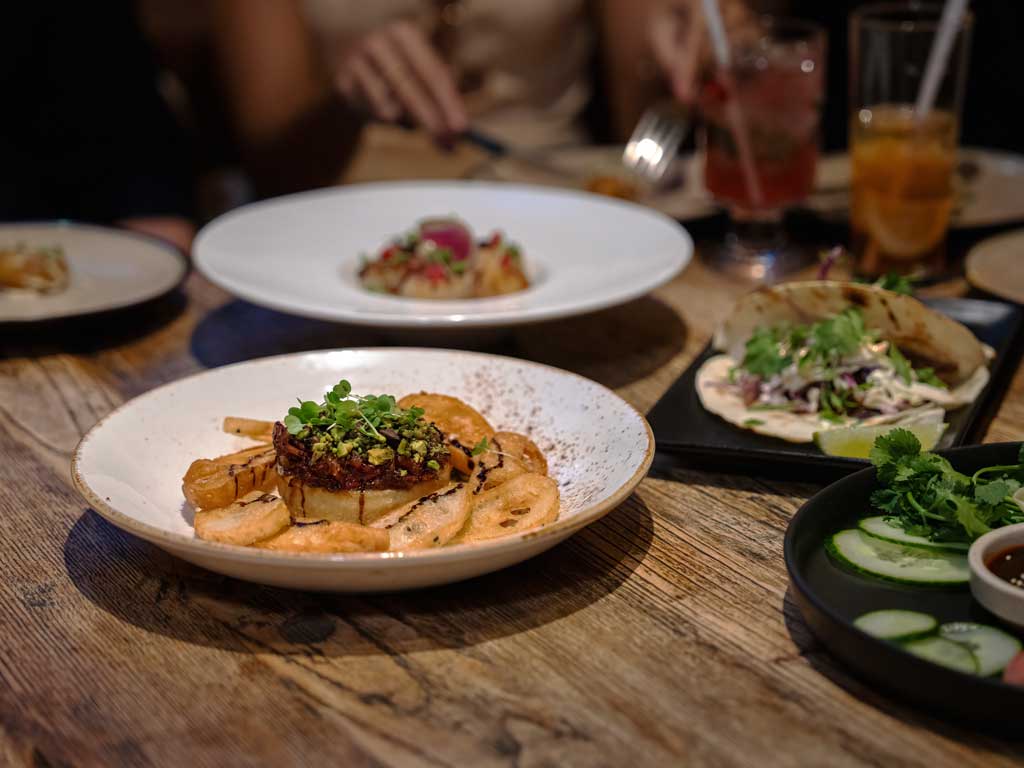 Share plates of food sit on a wooden table while a woman eats in the background