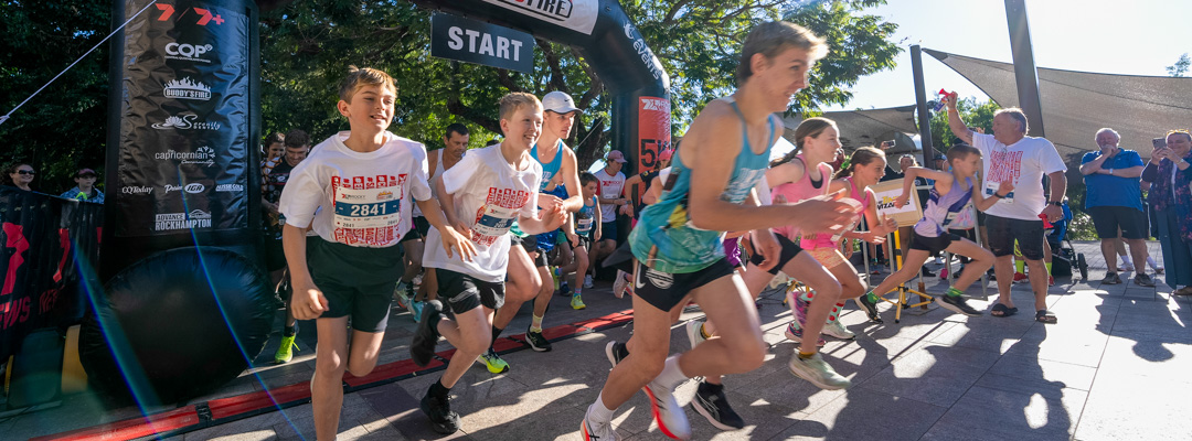 A group of children begins a running race under an inflated start line arch, as a man in the background uses an airhorn to start the race