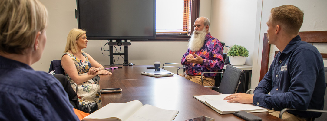 Four people sit around a wooden table having a discussion inside a conference room, with notebooks open in front of them
