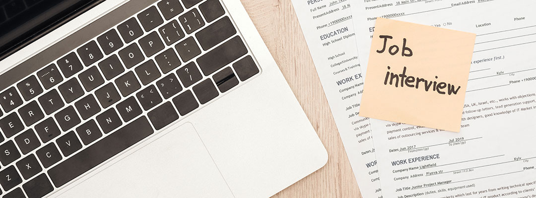 A laptop sits on a desk beside a resume, with a sticky note reading 'job interview'