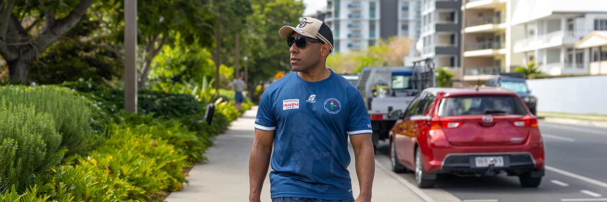 A man walks along a street, surrounded by green trees and buildings