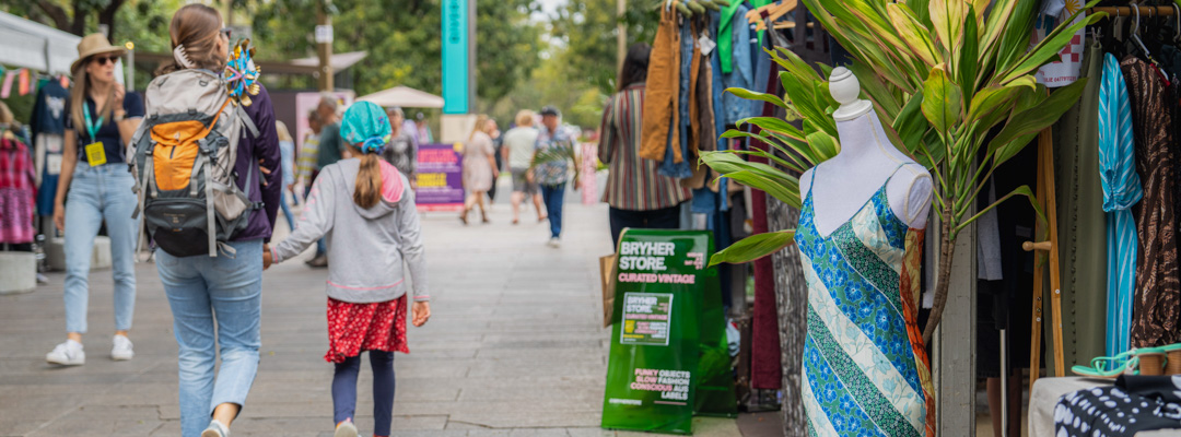 A mother and daughter walk hand-in-hand along a stone pathway, looking at market stalls selling clothing