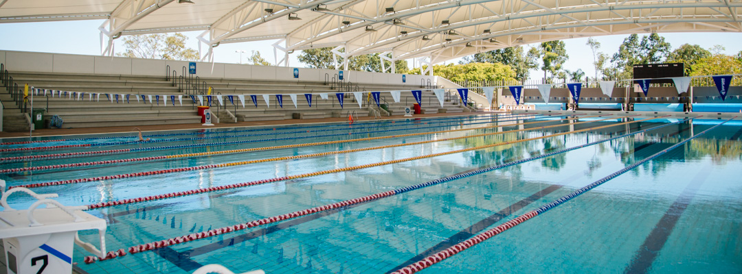 A large swimming pool marked with lanes and filled with blue water. Spectactor stands are in the background, and the pool is under a large shade structure