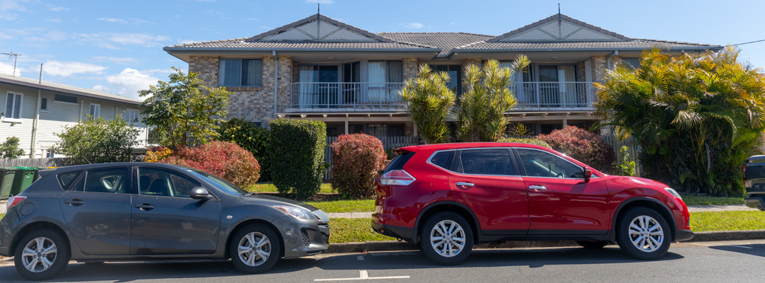 Two cars are parked outside a two-story brick duplex building surrounded by palm trees
