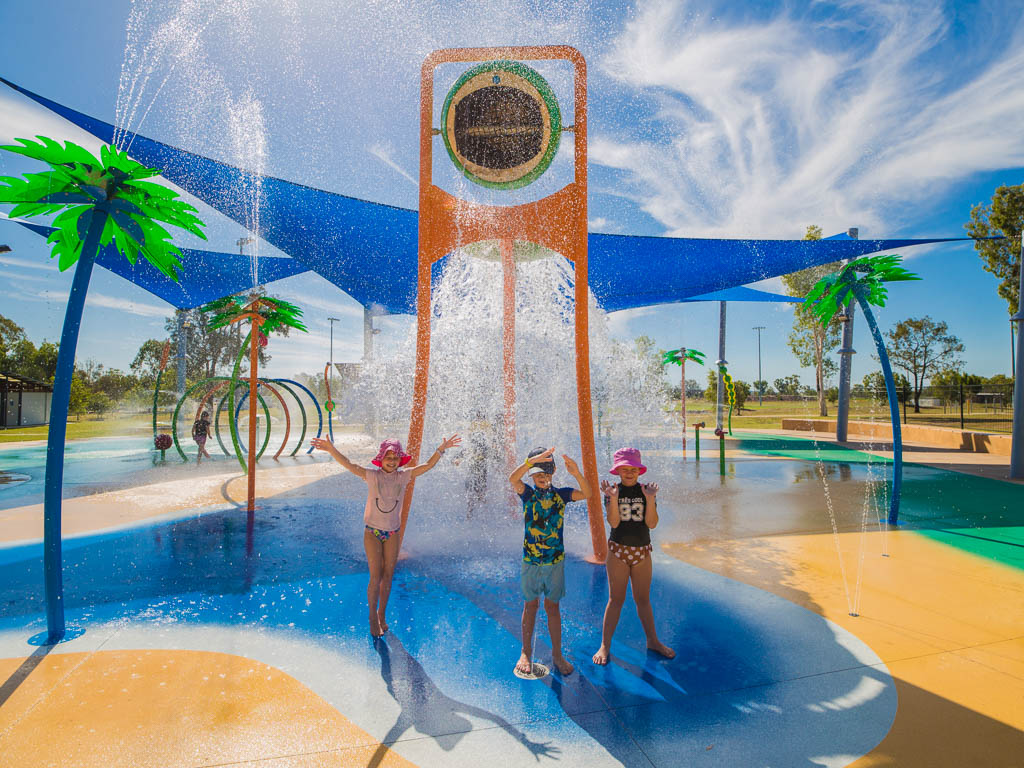 Three children are being splashed by water poured from a large bucket held up by a colourful structure at a water play area.