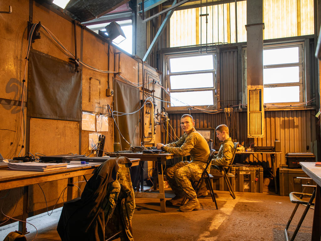 Two soldiers dressed in camoflagued clothing sit at a table inside an orange lid industrial building
