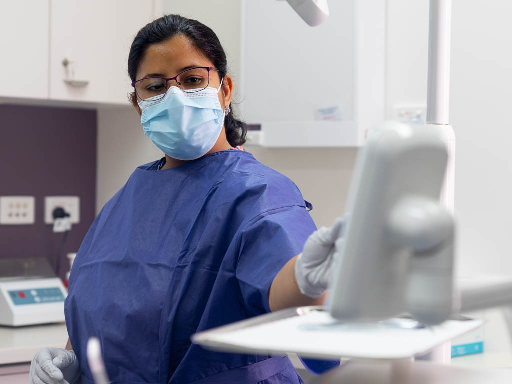 A woman wearing scrubs and a face mask looks at a screen built into a dental chair, while picking up dental equipment. A patient reclines on the chair, also looking at the screen.