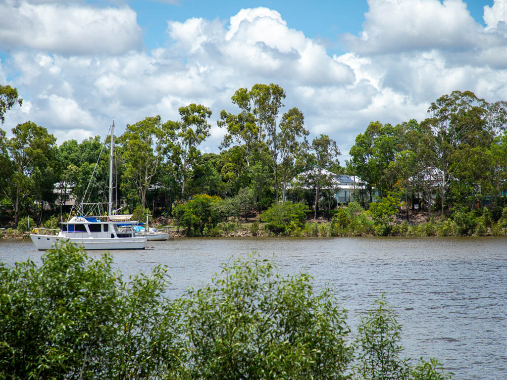 Looking over a blue river, where a row of houses sit behind trees on the other side of the water. A sailing boat is anchored in the river.