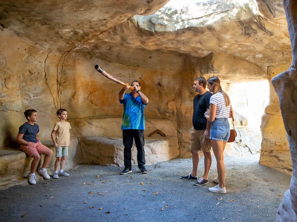 A mother, father and their two children watch on as a man plays the didgeridoo inside an artificial cave structure