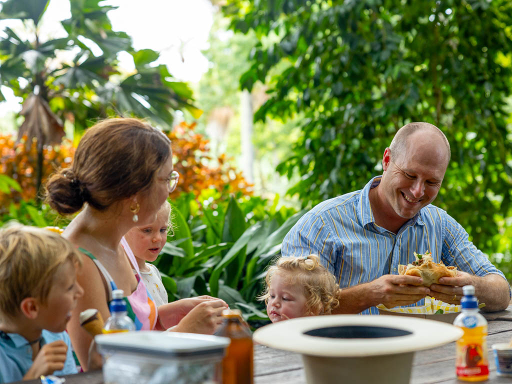 A father smiles as he eats a burger while sitting at a picnic table with his wife and children