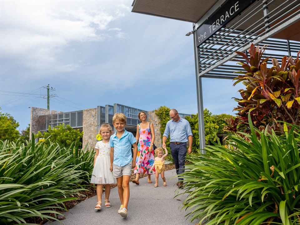 A young boy and girl hold hands as they walk along the footpath leading to a shopping centre, while their parents and sister follow behind