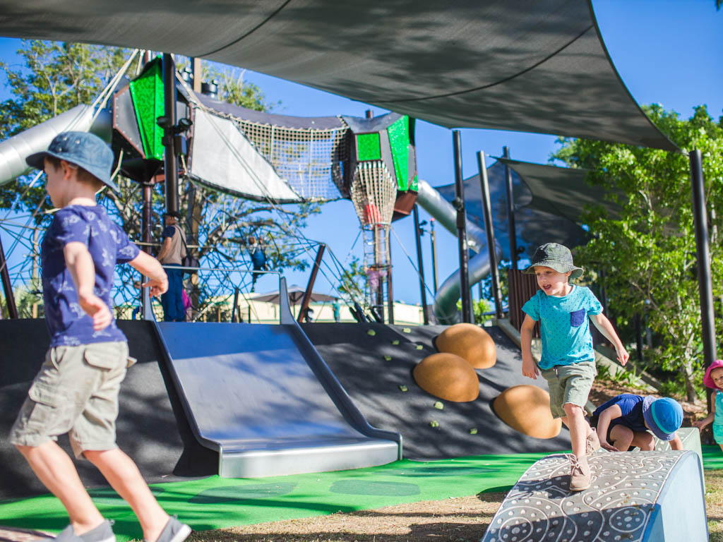 Two children are running across play-equipment ramps, in front of a large playground with slides, climbing walls and rope climbs