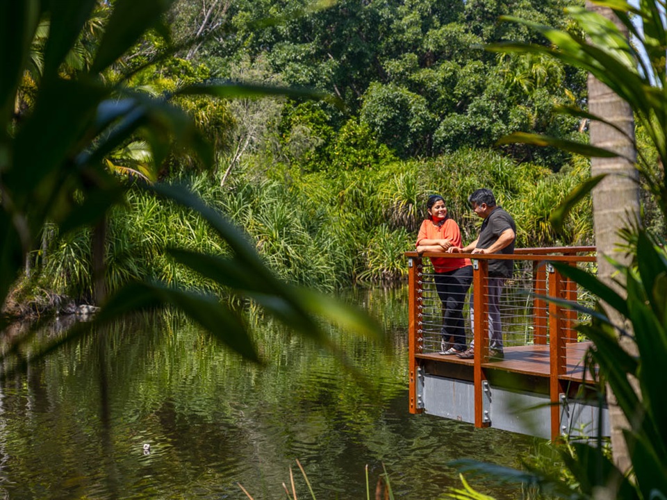 A woman and man are standing on a wooden platform, happily looking at each other, overlooking a lake surrounded by green trees and vegetation.