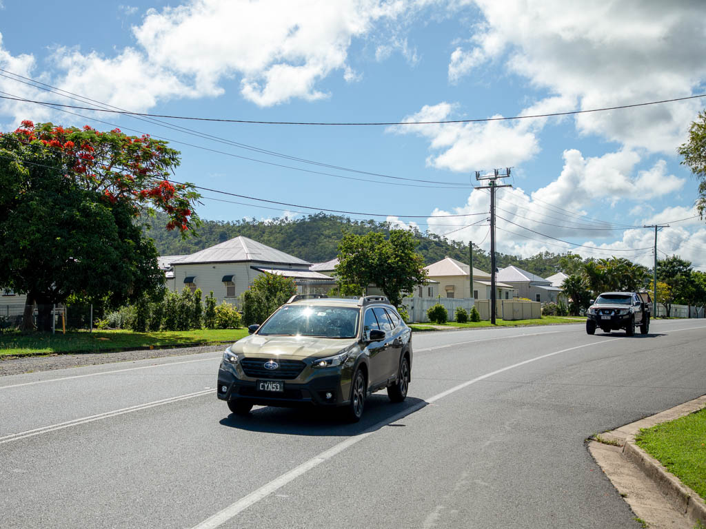Two cars pass by a row of cottage-style houses with mountains visible in the distance