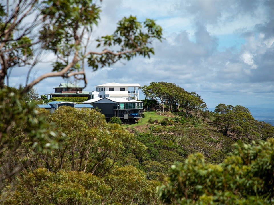 Large modern houses are perched on top of a hill, surrounded by vegetation and looking over a valley