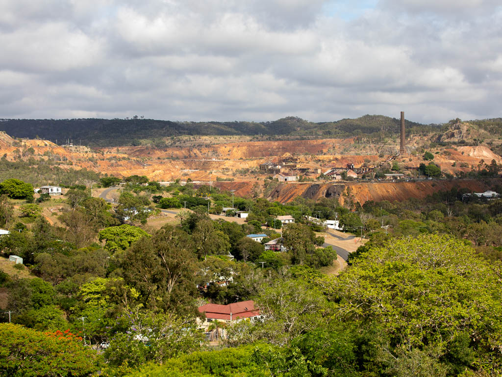 Cottage-style houses are nesstled amongst tall trees along a winding rode. In the background, the historic Mount Morgan mine site can be seen, dominated by a tall brick chimney