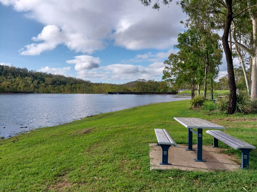 A shaded picnic table on green grass overlooks a blue lake with a dam wall off in the distance