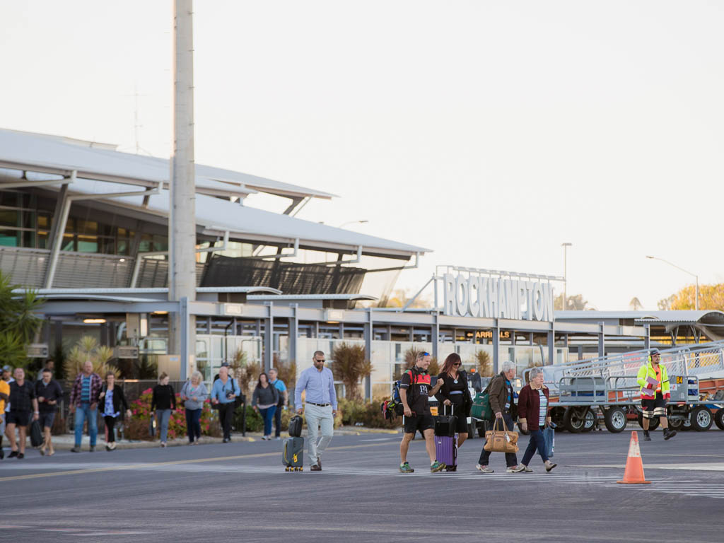 A row of people carry suitcases and bags cross the tarmac at an airport. The airport terminal is in the background, with the word 'Rockhampton' constructed on the roof