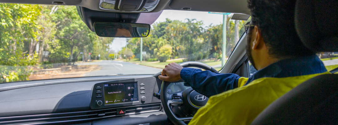 A man wearing high-visibility clothing sits inside his car, driving along a tree-lined street