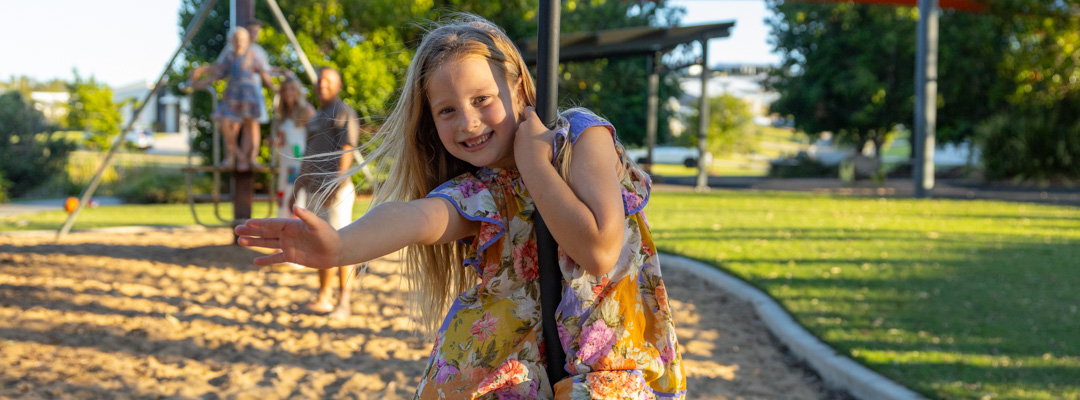 A young girl waves at the camera as she plays on a flying fox and her family watch from the background