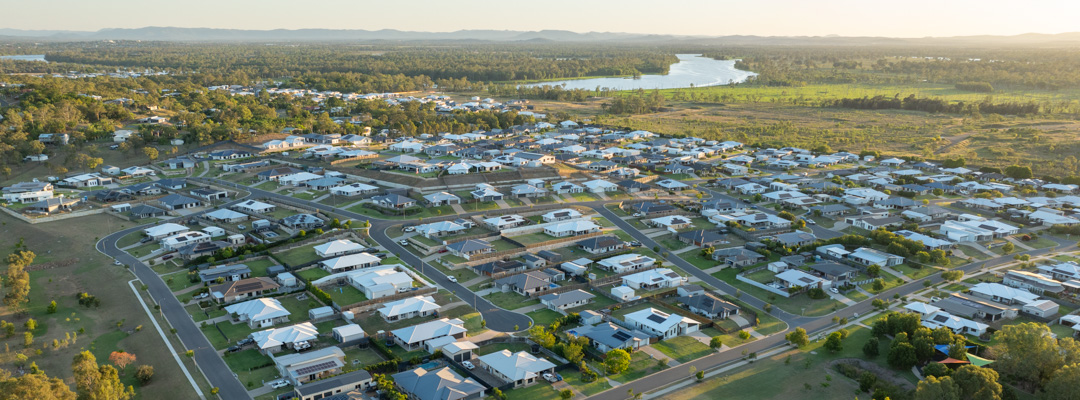 An aerial shot shows a new residential development, with streets of houses recently completed surrounded by green grass and trees backing onto a blue river
