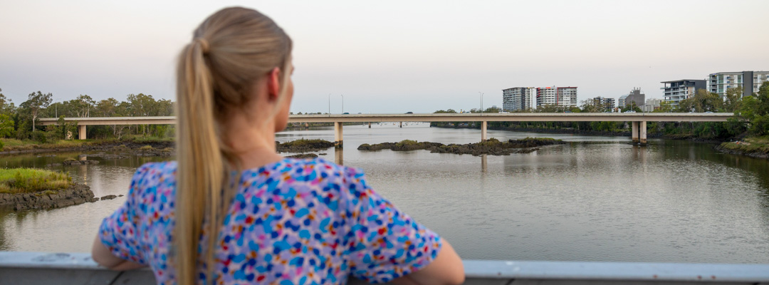 A woman leans against the railing of a bridge, as she looks across the Fitzroy River towards apartment buildings in Rockhampton's CBD. Cars can be seen travelling across a bridge in the distance