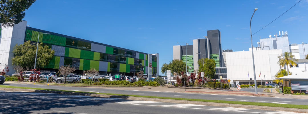 The large white frontage of Rockhampton Hospital sits beside a green and black extension. Cars are parked to the side and in a multi-level green carpark.