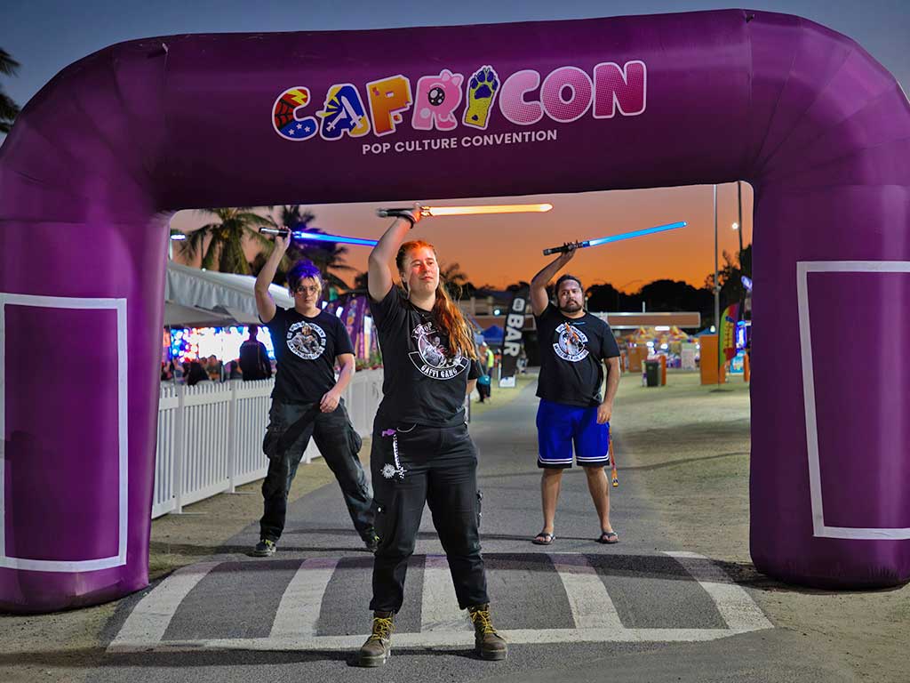 Three people swing illuminated swords in the area under an arch with the CapriCon logo on it'