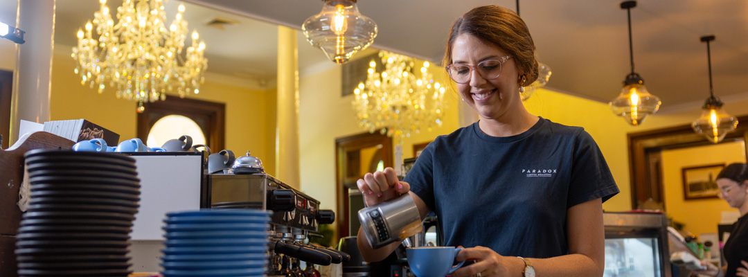 A cafe worker smiles as she pours milk into a coffee up